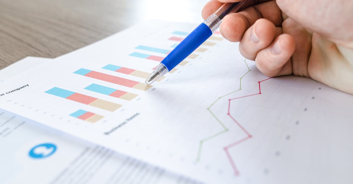 A close-up of a hand with a pen analyzing data on colorful bar and line charts on paper.