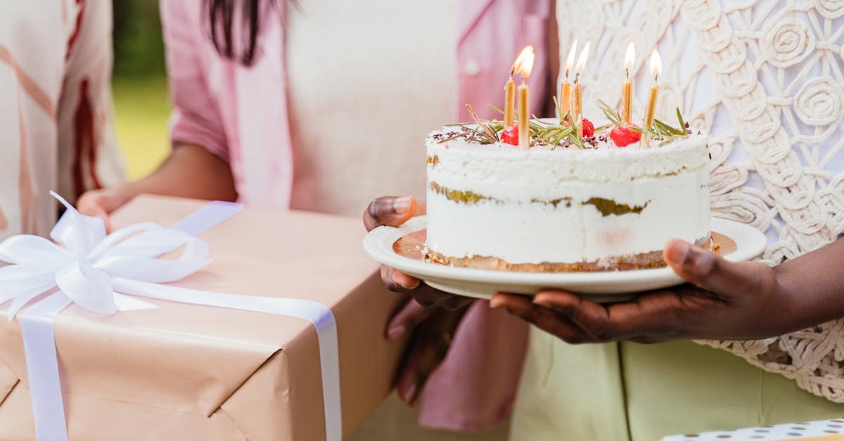 Close-up of a birthday cake with lit candles and a wrapped gift held by women during a celebration.