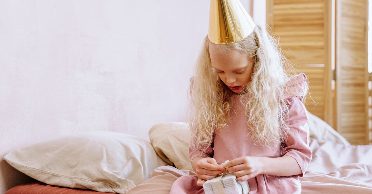 A young girl sits on a bed opening a gift while wearing a party hat, celebrating her birthday.