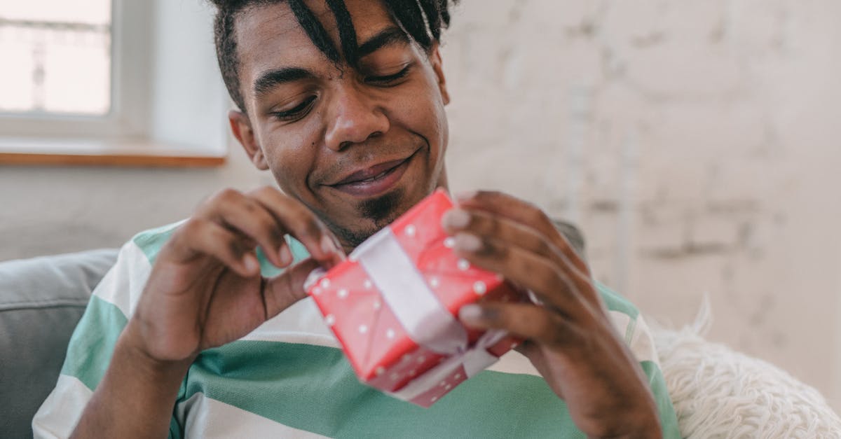 A joyful man with dreadlocks happily opens a red gift box while sitting on a couch.