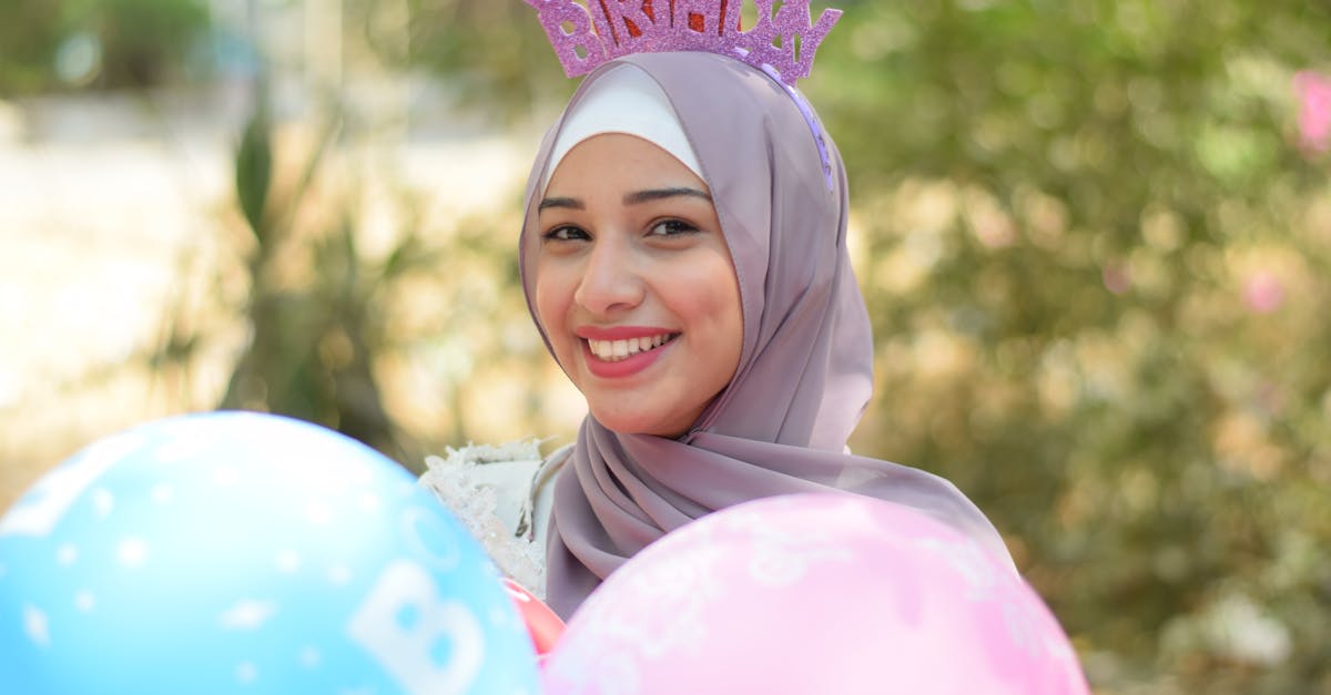 Smiling woman wearing hijab and birthday hat, holding colorful balloons outdoors, celebrating a joyful birthday.