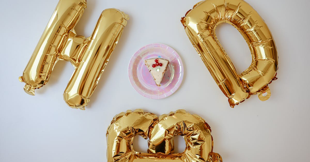 A festive birthday setup with a cake slice and shiny gold foil balloons spelling 'HBD'.