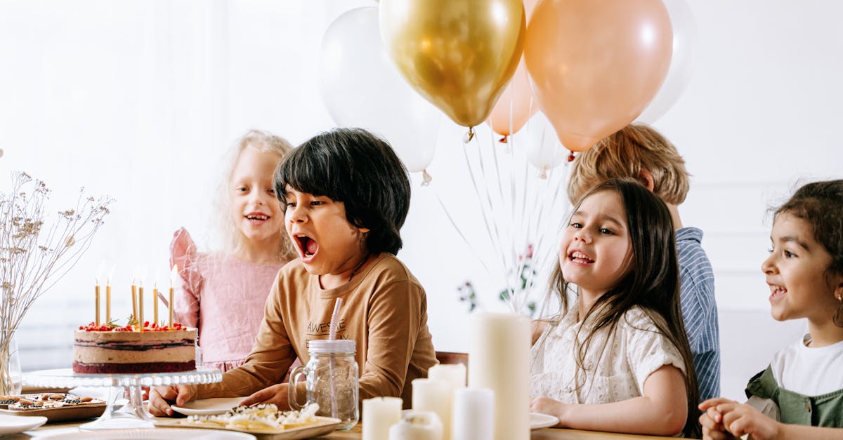 Children celebrate a birthday, blowing candles on a cake with balloons and decorations indoors.