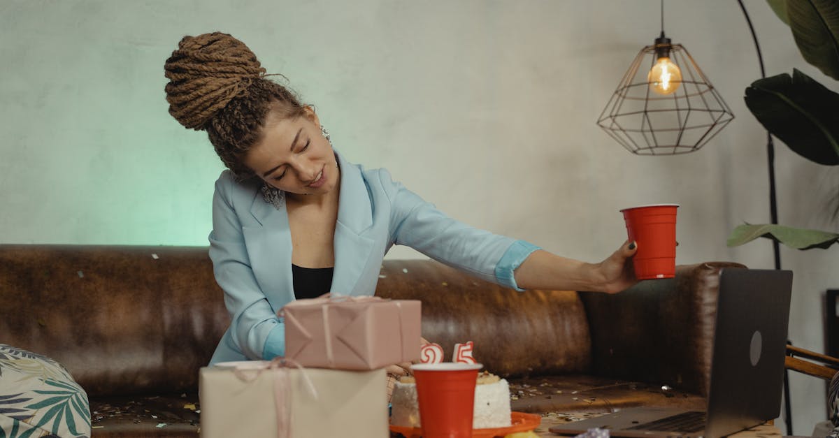 A woman enjoys a virtual birthday party with cake, gifts, and a video call setup.