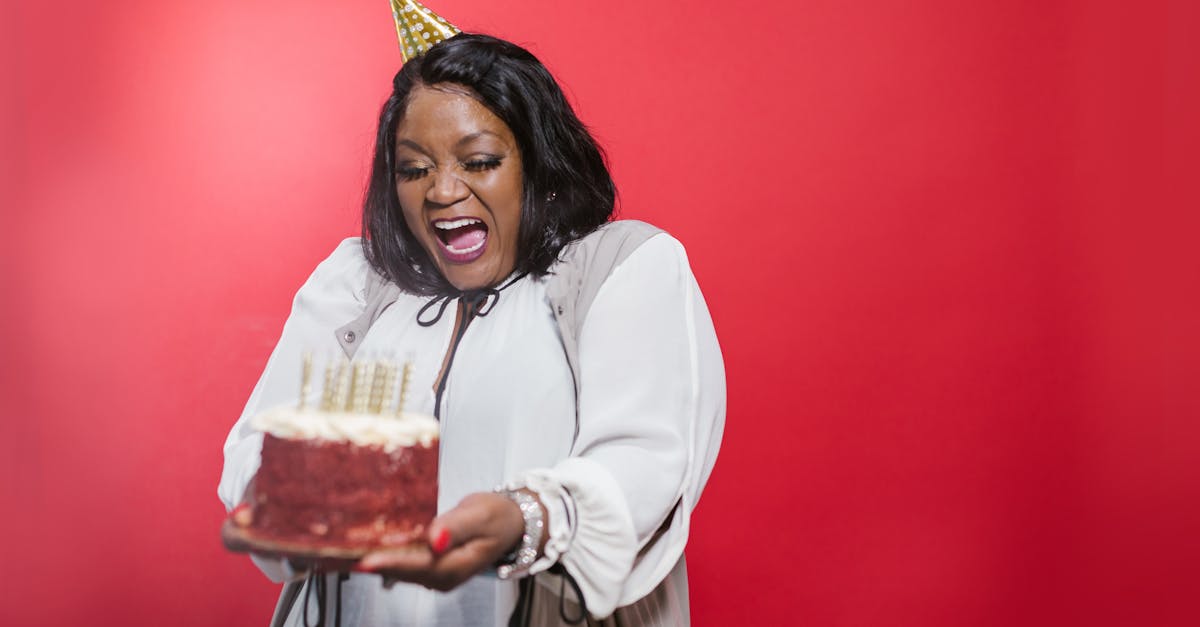 A joyful woman wearing a party hat holds a birthday cake against a vibrant red background.